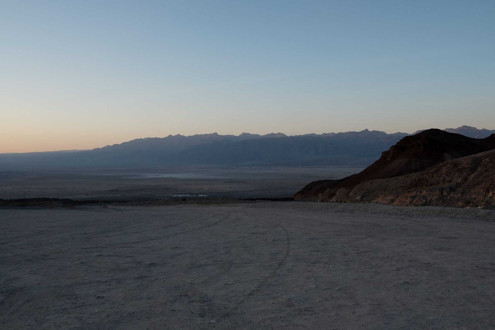 Flat landscape at sunset, taken from a bare dirt parking lot. Mountains in the background fade into the sunset